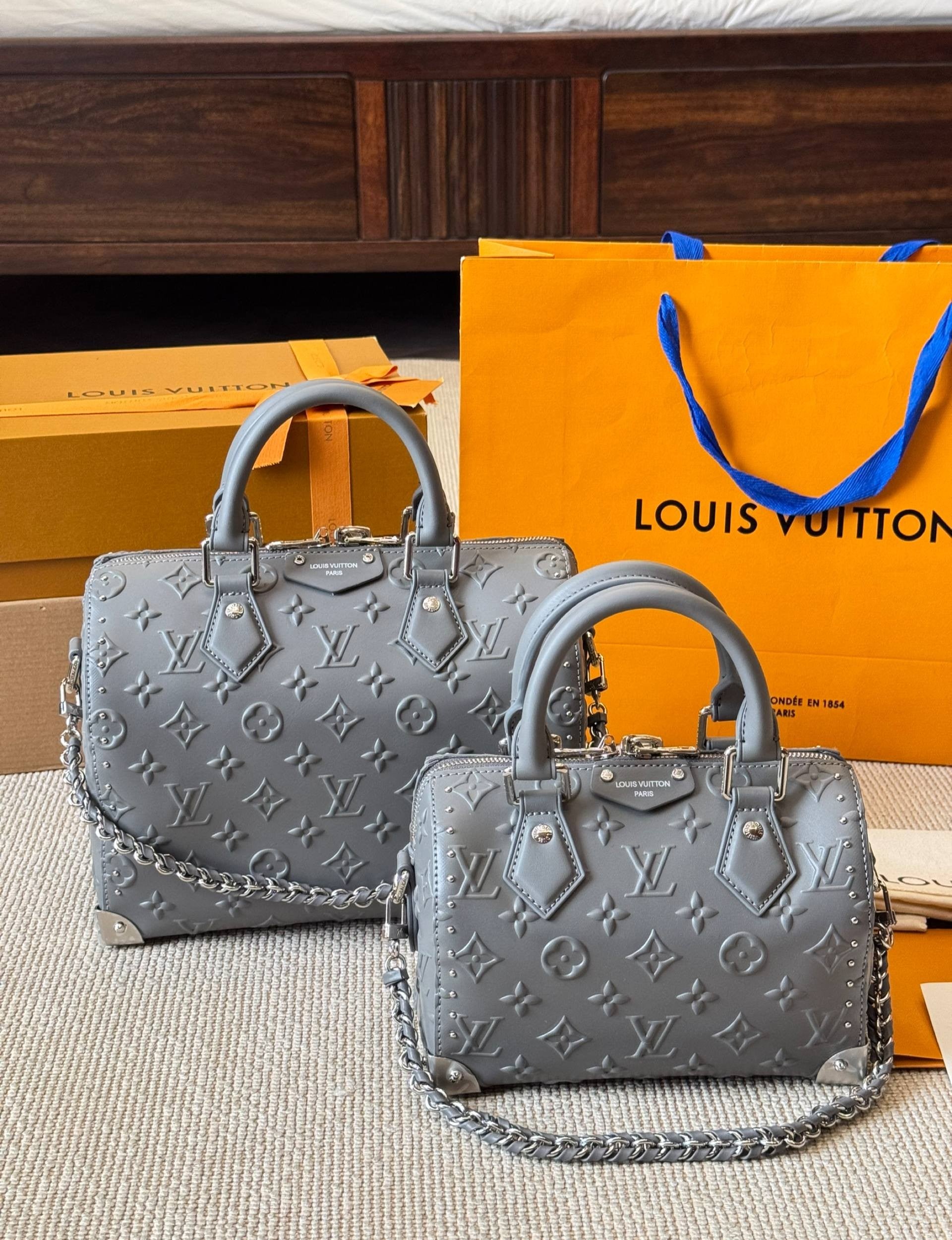 Two gray handbags with a Louis Vuitton logo on a wooden floor with an orange Louis Vuitton bag in the background.