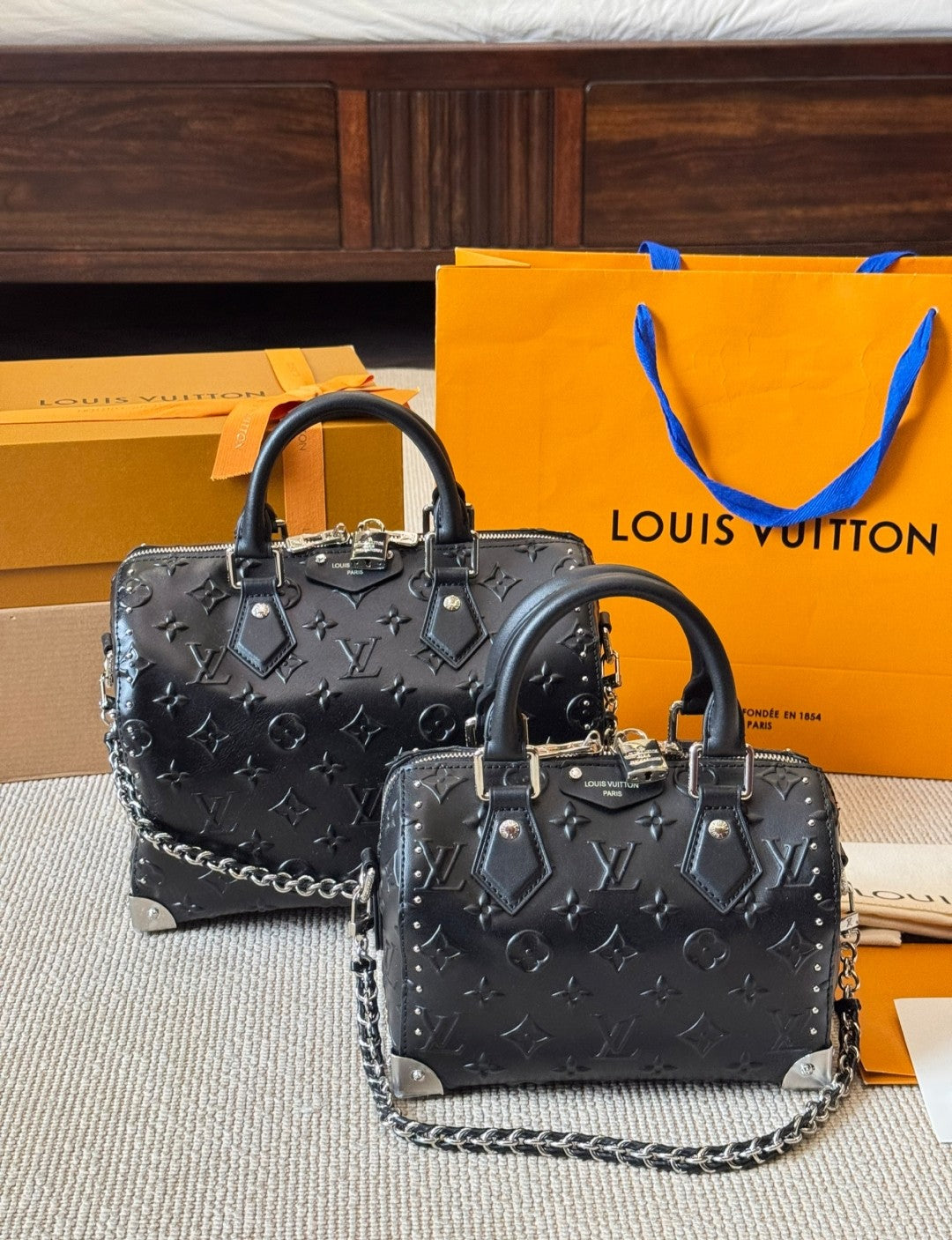 Two black handbags with silver chains on a surface with a Louis Vuitton orange bag and box in the background.