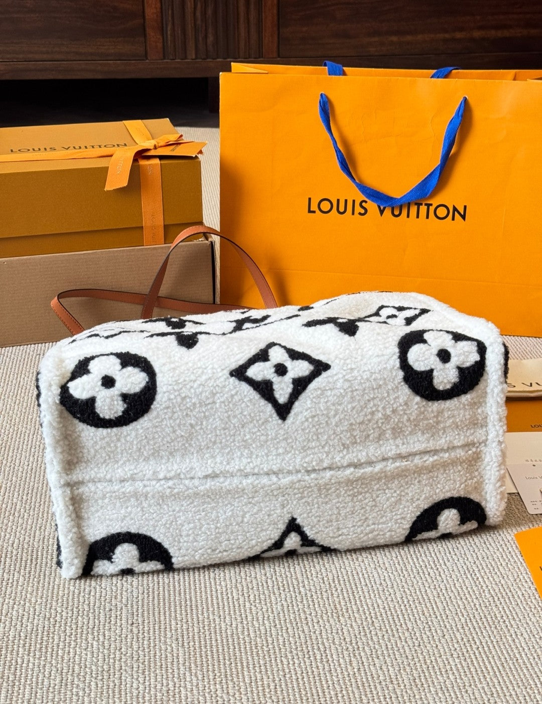 White pouch with black floral patterns on a surface with a Louis Vuitton shopping bag in the background.