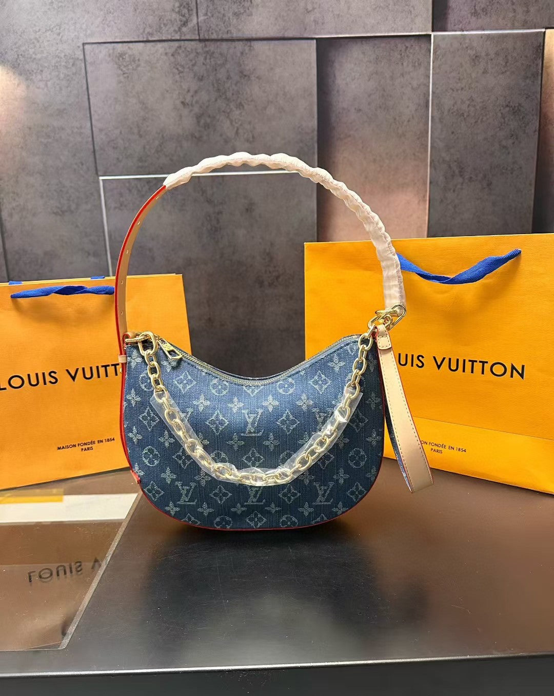 Blue handbag with gold chain detail on a reflective surface with Louis Vuitton packaging in the background.