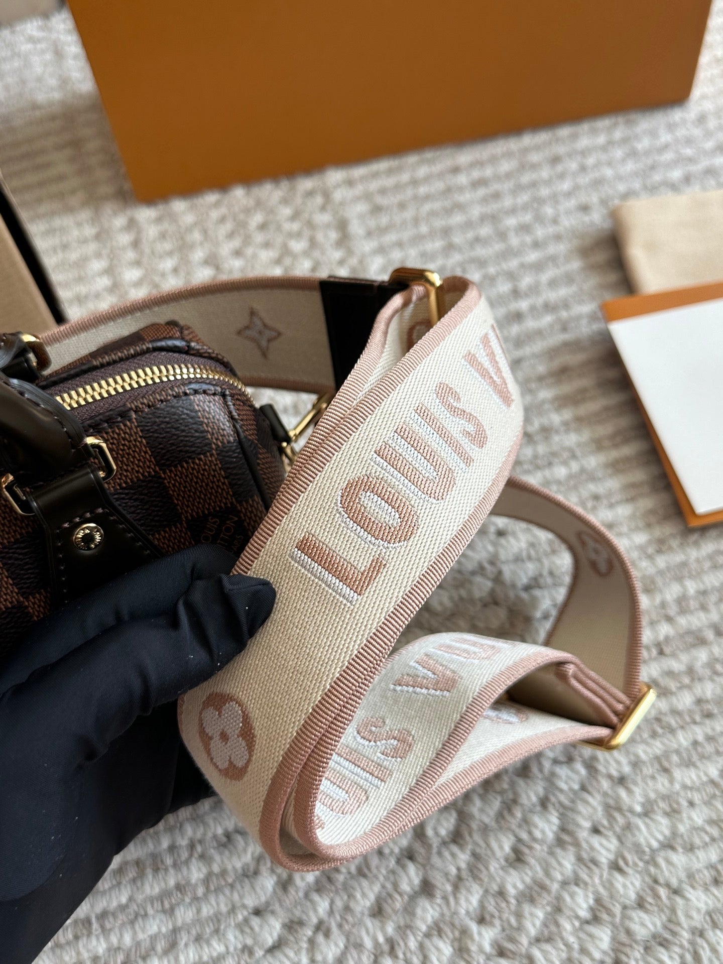 Louis Vuitton belt with monogram pattern held by a gloved hand, with a part of a Louis Vuitton bag and box in the background.